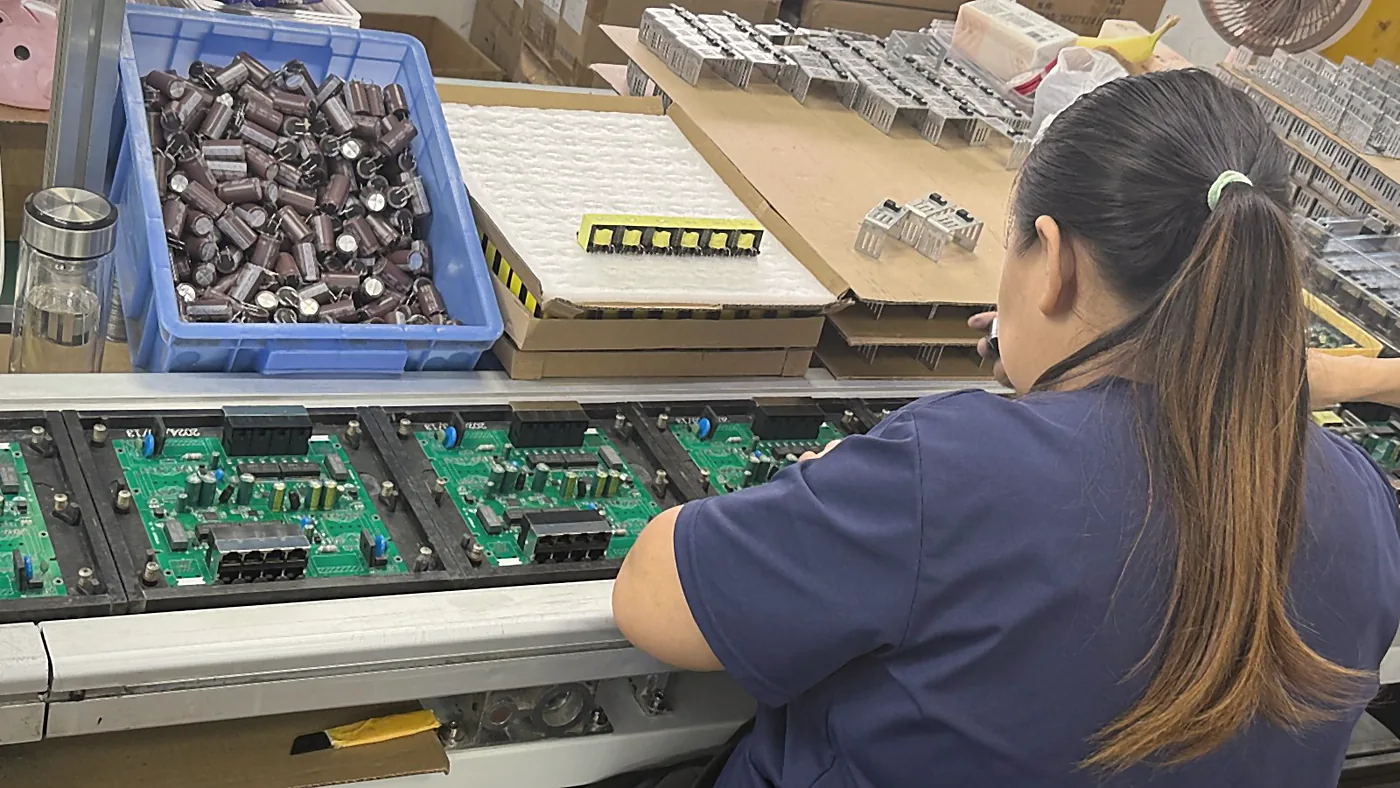 An operator at an assembly line, circuit boards and a box of electrolytic capacitors in front of her